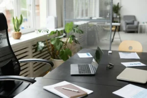 Biurko duże w jasnym home office: czarny blat z laptopem, notatnikiem i okularami; ergonomiczne krzesło i rośliny przy oknie.