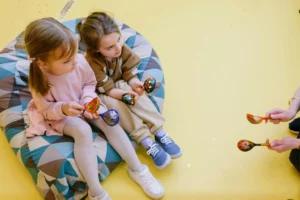 Dwie małe dziewczynki wygodnie siedzą razem na dużej, wzorzystej pufy dla dzieci i bawią się kolorowymi, drewnianymi łyżkami na żółtym tle.